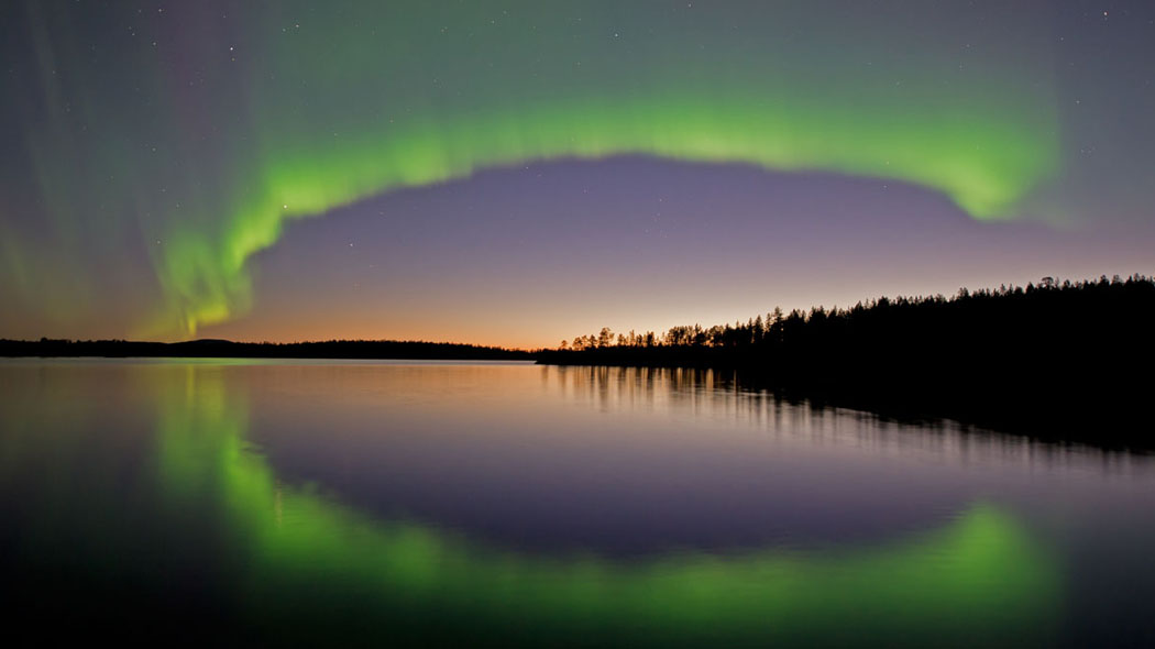 Grönt norrsken lyser upp himlen, färgerna reflekteras i den mörka vattenytan. En mörk skog delar himlen och vattnet i horisonten.