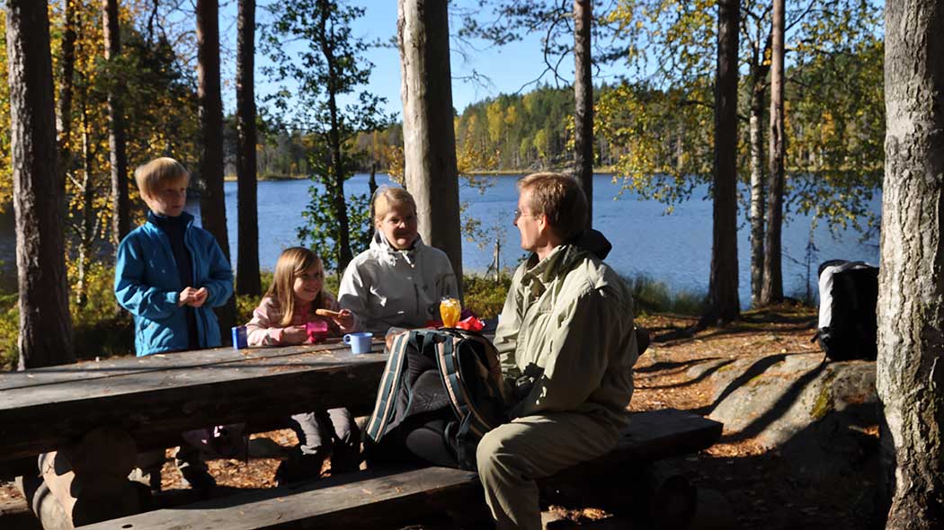 En familj vid en bord i ett höstskogslandskap vid en sjö.