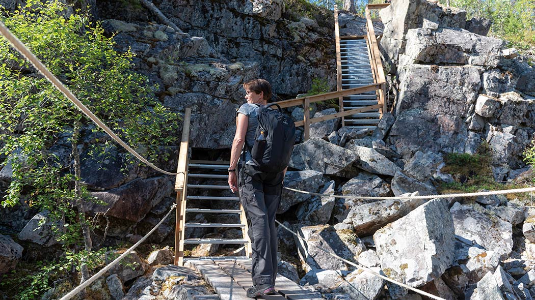 En vandrare står med ryggsäcken på ryggen i nedre änden av trappor som klättrar uppför en brant stenig sluttning. Ett repräcke leder till trappor, och trappor har träräcker.