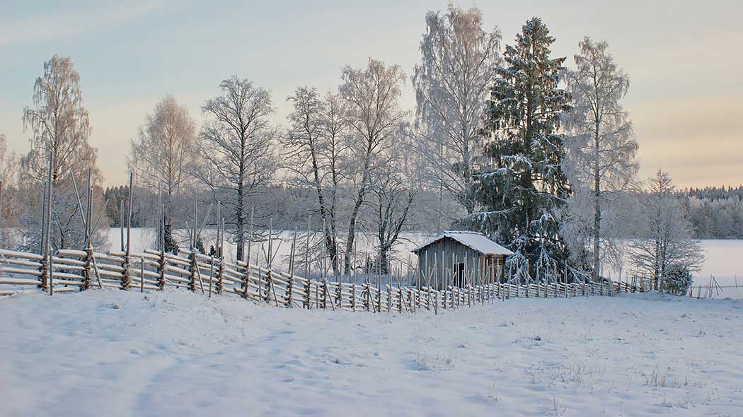 Snölandskap med fält och staket i förgrunden. I bakgrunden finns en gammal byggnad, träd och en snöig sjö.