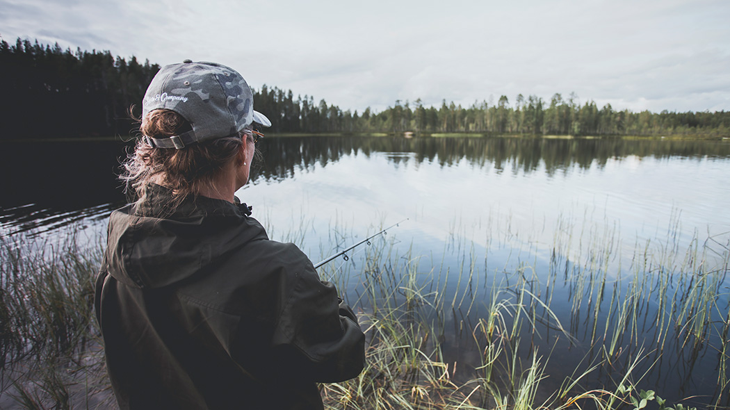 En vandrare som fiskar i Kylmäluoma. I bakgrunden ett lugnt sommar sjölandskap.