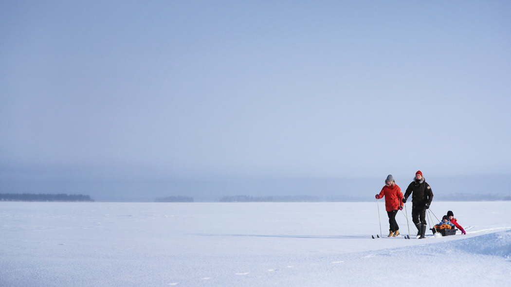 Två vuxna åker skidor på en snöig och öppen sjö. Den andra följs av en ackja med två barn.