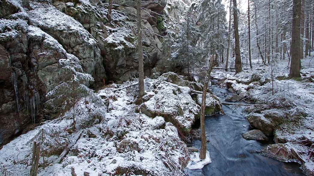 En bäck rinner i skogen, bredvid ett berg. Det ligger ett tunt snötäcke på marken.