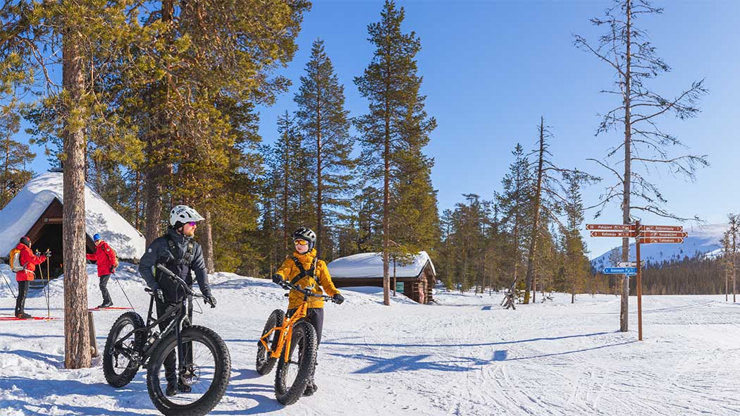 I förgrunden finns två personer som lutar sig mot svarta fat bike-cyklar i snödrivan. En kåta och ett litet vedlider kan ses i bakgrunden i det vintriga landskapet. En tallskog och en blå himmel kan ses i omgivningen.
