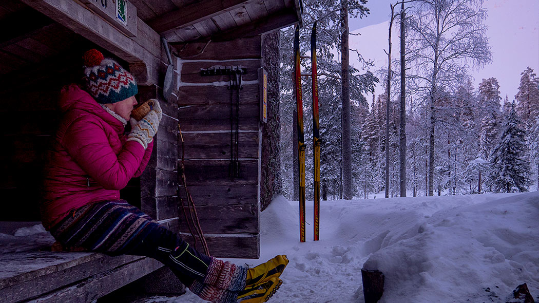 En kvinna sitter i skärmskyddet och dricker något hätt. Skidor står i snön. Vinterfjälllandskap i backgrundet.