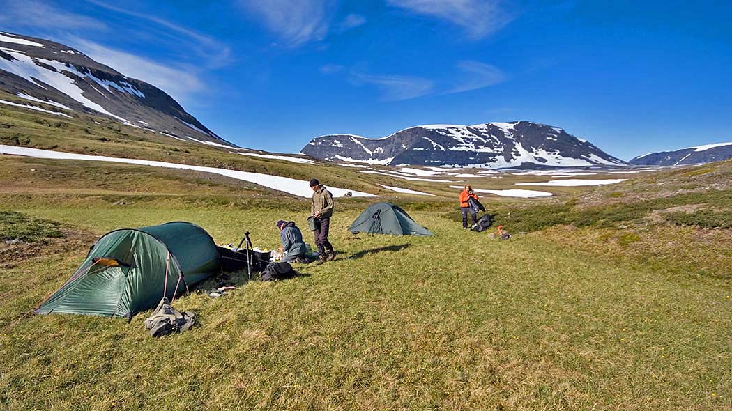Tre personer som sätter upp tält i öppen bergterräng i soligt väder. Höga bergtoppar i bakgrunden.