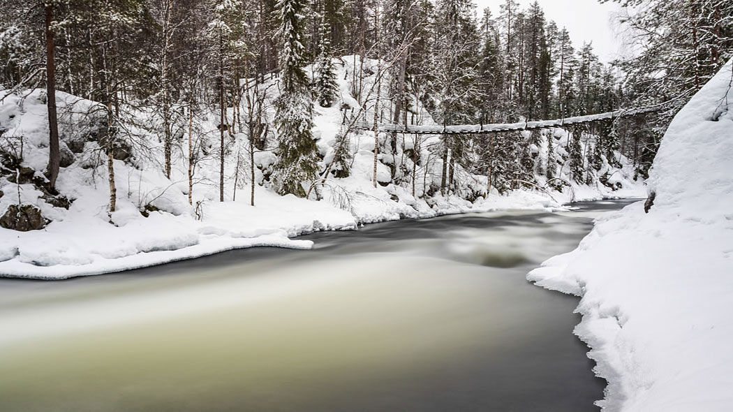 Ett snöigt landskap med en hängbro ovanför en strömmande flod.