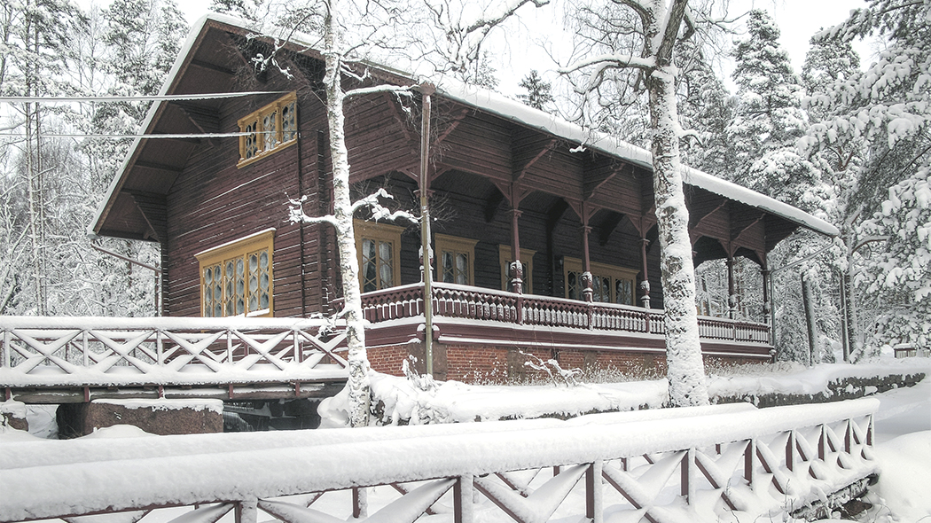 I vinterlandskapet en mörk träbyggnad med en stor veranda. Några träd framför byggnaden och en snöig skog bakom.