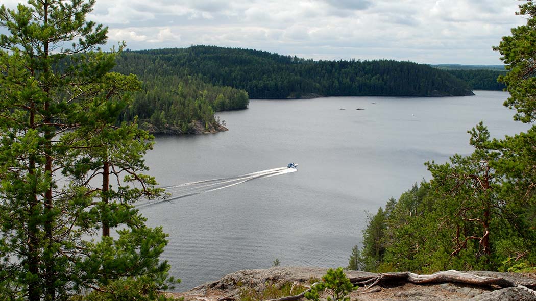 Ett sjölandskap öppnar sig, sett från toppen av fornborg. På den somriga sjön finns en motorbåt.