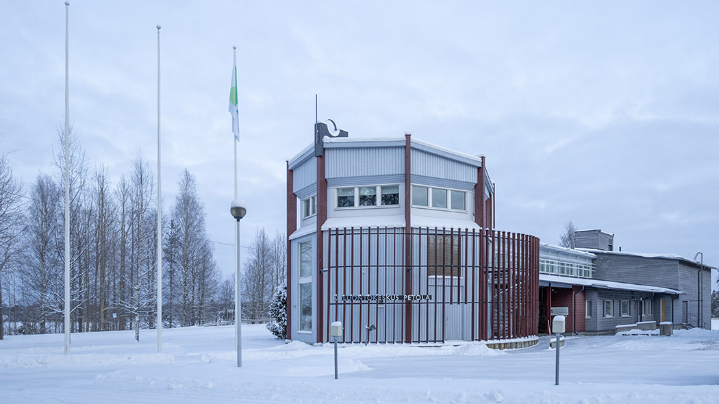 En stor byggnad i ett vinterlandskap. I förgrunden tre flaggstänger, varav en har flagga. Det finns en snöig sjö i bakgrunden.
