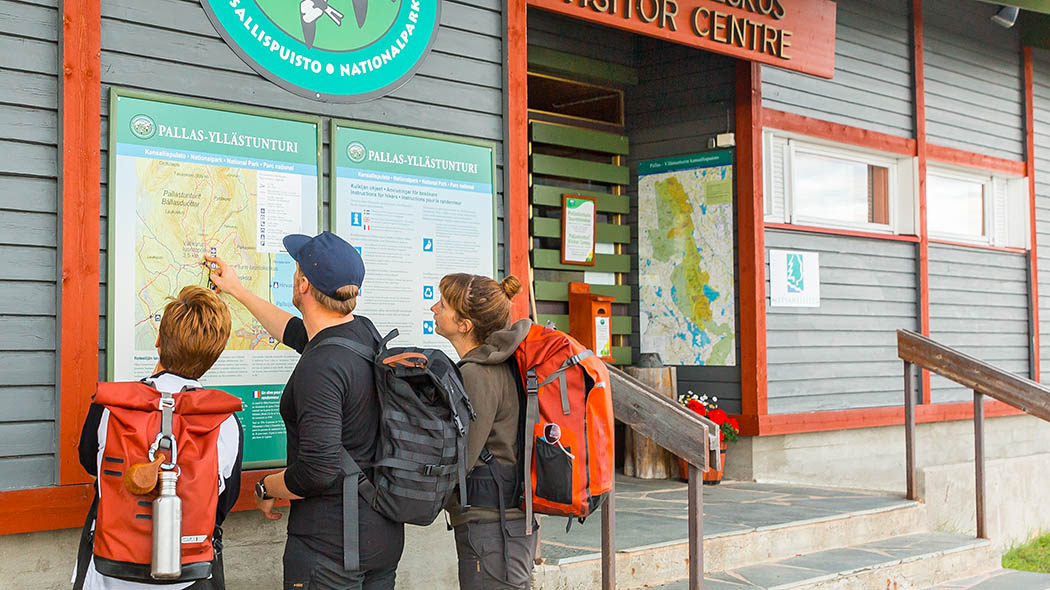 På den yttre väggen bredvid naturumets ytterdörr finns nationalparkens emblem, en informationstavla och en karta. Tre vandrare tittar på Pallas-Yllästunturi kartan som finns på väggen.