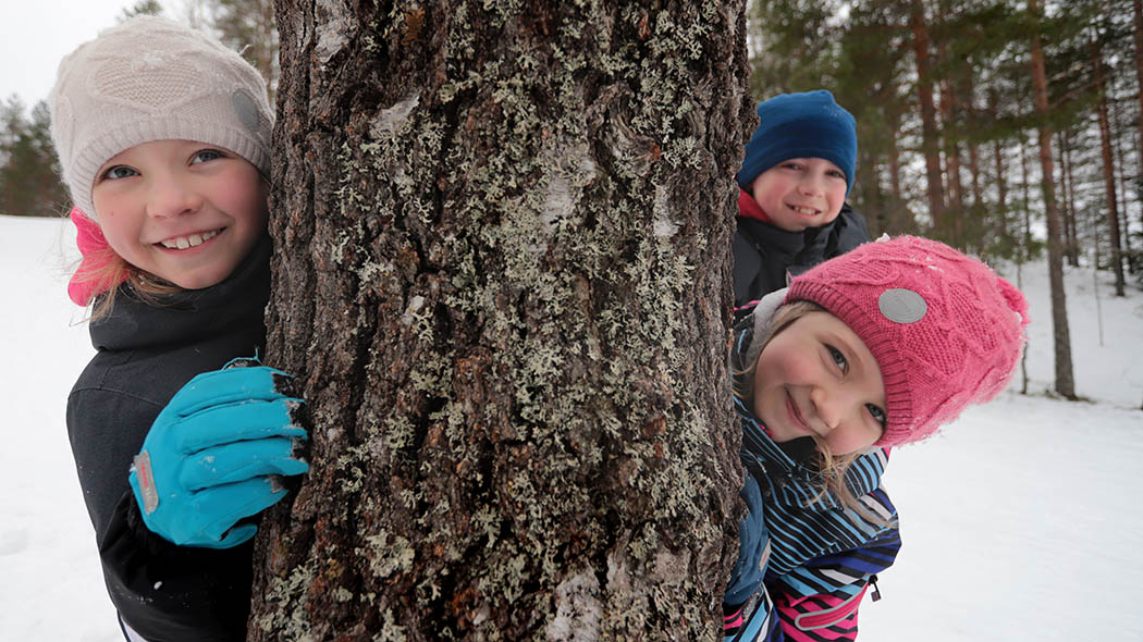 Tre barn tittar bakom ett träd. Det ligger snö på marken.