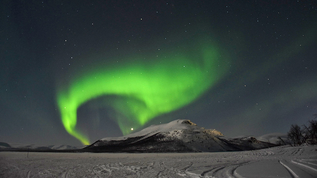 Ett vinterlandskap med ett fjäll bakom en frusen sjö. Norrskenen och stjärnor lyser på himlen.