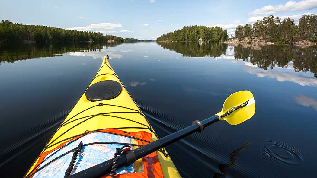 I förgrunden på bilden finns en paddel och en kanot med en karta fäst vid dess däck. Det finns skogar och klippor vid sjön.