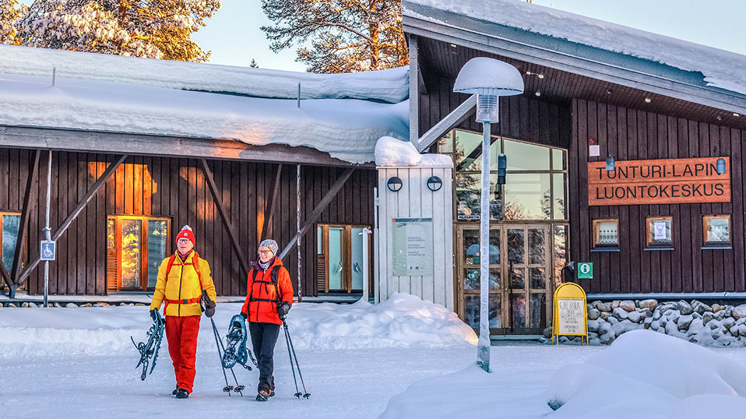 Två vandrare går framför huset på vintern. De bär snöskor och stavar.