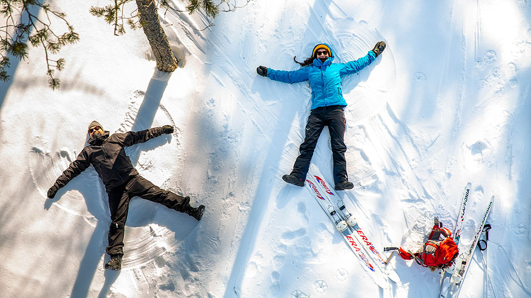 En kvinna och en man ligger på en snöboll och gör snöänglar. Bredvid dem står skidor och en ryggsäck.