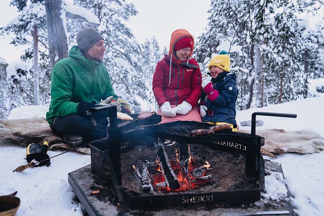 Två vuxna och ett barn sitter på en renhjord vid en eldplats på vintern.